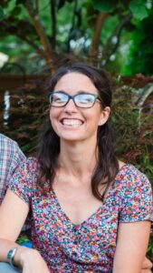 Screenshot - Drayton Avenue Cooperative Preschool Smiling woman with dark hair and blue glasses outdoors, wearing a colorful floral dress.